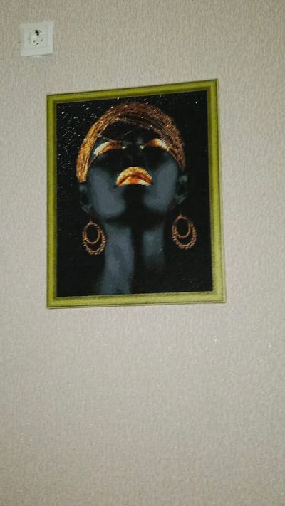 A framed artwork depicting a woman's face, painted in dark tones with gold accents, hangs on a textured, light-colored wall. The artwork is square and bordered by a thin, light green frame. The woman's eyes are closed, and her lips are parted, painted in a metallic gold. She wears gold hoop earrings and a gold headpiece. To the left of the artwork, a white electrical outlet is mounted on the wall.