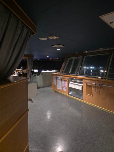 The image shows the interior of a ship's bridge at night, likely docked in Brindisi, Italy. The bridge is spacious with large windows offering a view of the dark water and distant lights of the city or harbor. The room is dimly lit, with the primary light sources appearing to be overhead ceiling lights and the reflected lights from outside.
The foreground is dominated by a wooden structure on the left, possibly a desk or console, with a grey curtain draped over it. The floor is a grey, speckled material. In the mid-ground, there is a control panel or console with various instruments and screens, some of which are illuminated. Behind this, a large white console-like structure is visible. More wooden paneling lines the walls, with metal railings and various notices or charts affixed to them. The ceiling is dark blue and appears to be made of acoustic tiles.
The overall impression is one of functional, utilitarian design, characteristic of a ship's bridge. The presence of nautical equipment and the view of the water suggest it is a working vessel. The darkness and the distant lights suggest it is late at night. The exact location within Brindisi is not discernible from the image itself, but the context points to a port or harbor setting. There are no people visible in the image.