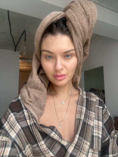 A young woman is centered in the image, wearing a brown towel wrapped around her head, and a brown and tan plaid shirt open at the neck. She has fair skin, blue eyes, and slightly parted lips. A silver necklace with two pendants, including a cross, is visible on her neck. A tattoo is slightly visible on her chest area.
The background shows a glimpse of a room interior, including a light fixture and a door. The photo appears to be taken indoors. There are no indications in the photo that would link it to Gavrilkovo, Russia.