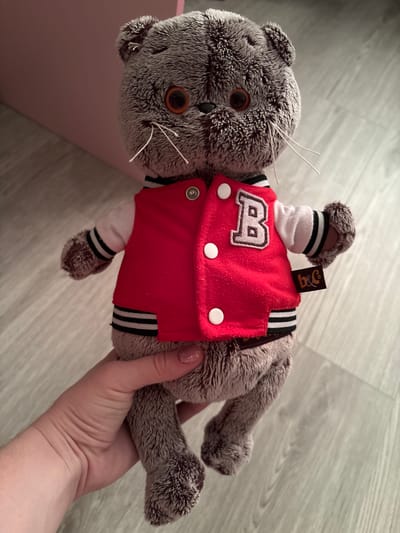In this medium shot, a grey, plush cat toy is held in a person's hand. The toy is lying on its back, with its head slightly tilted to the left. It is wearing a red jacket with white trim and a white "B" emblem. The background is a light-colored wooden floor. The image appears to be taken indoors during the daytime, with artificial lighting. The person's hand is visible at the bottom of the frame, gently supporting the toy. There is a pink object partially visible in the lower left corner. A small, dark brown tag with the letters "B&C" in gold is attached to the toy's neck.