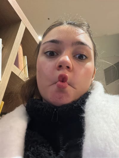 A woman poses with pursed lips, looking directly at the camera from a low angle. She's wearing a black jacket with white fur accents and visible zipper. A shelf is partially visible in the lower left corner, cluttered with books. The background is blurred, giving a sense of being indoors with bright, diffuse lighting.