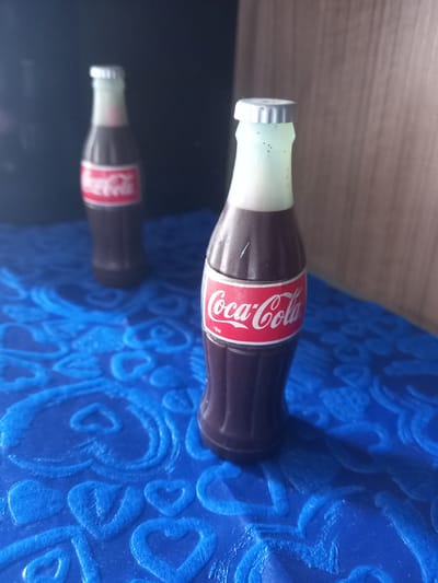 A slightly angled close-up shot depicts a Coca-Cola bottle resting on a shiny surface that reflects the bottle. The bottle has a silver cap, and the neck area beneath the cap is covered in a light-green coating. The Coca-Cola logo is prominently displayed on a red band around the bottle. The bottle is on a blue surface with raised patterns resembling hearts or stylized floral designs. The setting appears to be indoors. The location is in the city of Trujillo, Peru.