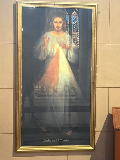 Framed print depicting the Divine Mercy of Jesus adorns a light-colored wall. Jesus is shown with rays of light emanating from his heart in red and white. The words "Jesús, en Ti Confío" are on the right. A stained glass window is visible in the upper-left corner of the image. The frame is gold-colored and rectangular. The wall appears to be made of tiles.