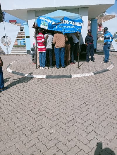 The image displays an outdoor scene in Mumbuni North ward, Kenya, under bright daylight with visible shadows. The ground is paved with a herringbone pattern of concrete pavers. A circular concrete island with black and white curbing is situated in the foreground. Centrally, a light blue canopy tent is erected, displaying text including "MACHAKOS TEACHER" and "ISO 9001:20...". Approximately 6-8 individuals, primarily men, are clustered closely around and within the tent's immediate vicinity, facing inwards, suggesting engagement with its contents or services. One man wears a red and white striped shirt and blue jeans, another a brown jacket and blue jeans. To the right of the tent, a female security officer in a dark blue uniform, including a cap and a badge, stands observing with her hands in her pockets. Adjacent to her, a man in a blue and white striped long-sleeve shirt, blue jeans, and a dark baseball cap also stands, observing the scene. In the background, a light blue and white building structure, possibly an entrance or gatehouse, is visible. To the left of this structure, a large white gate with a distinct circular design is partially visible. Additional buildings are present in the distance beyond the gate. The lower body of another individual is partially visible on the far left of the frame.