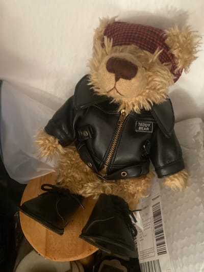 A stuffed teddy bear is positioned upright, wearing a black leather jacket with a "TEDDY BEAR" patch and a red and black checkered cap. The bear is sitting on a circular wooden object, with two black lace-up boots placed in front of it. The bear is situated within a white box, with a white bubbly packing material and a shipping label visible in the background. The shipping label contains German text and a barcode. The overall scene suggests the teddy bear is either being shipped or has recently arrived, possibly to or from Kassel, Germany, given the German text on the label. There are no people present in the image.