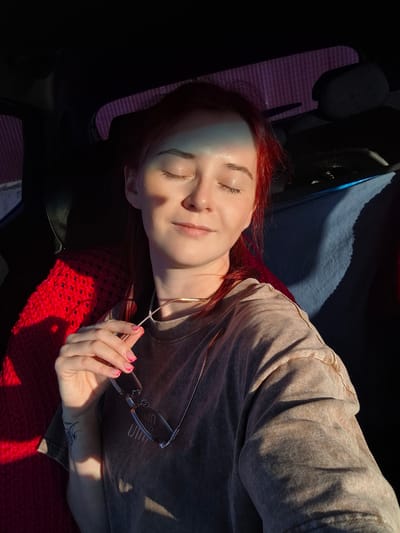 A young woman with red hair is sitting in the passenger seat of a car, looking towards the right with her eyes closed. Sunlight is illuminating her face, casting a shadow on the left side of her face and across her shirt. She is holding a pair of glasses in her right hand, and has a tattoo on her left forearm. A red knitted blanket is draped over the seat behind her. The background of the car interior is dark, with a textured surface visible through the back window, suggesting a location in Kamyennoye, Russia.