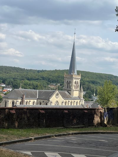 This image captures a view of the Église Saint-Martin in Le Houlme, France. The church, constructed from light-colored stone, features a prominent tall, grey spire topped with a cross. Its facade exhibits Gothic architectural elements, including arched windows and decorative stonework. The church is situated amidst a landscape of lush green trees and rolling hills, suggesting a semi-rural setting within or on the outskirts of the town. In the foreground, a low, weathered brick wall runs horizontally across the frame, partially obscuring the view of the church. Beyond the wall, a paved parking lot with white parking lines is visible. The sky above is partly cloudy, with patches of blue peeking through. The overall impression is one of a historic religious building set against a natural, verdant backdrop.