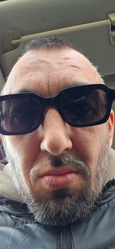 A close-up portrait captures a man with his head tilted significantly to the left within the frame, showing him from a slightly upward angle. He wears dark, square-framed sunglasses, which obscure his eyes. His face is characterized by a full beard and mustache, heavily streaked with grey and white hairs, giving him a salt-and-pepper appearance. His lips are slightly parted, revealing a neutral expression.
He is dressed in a dark-colored jacket, possibly a padded or puffer style, with a visible zipper detail on the left side of the image. The skin on his face shows visible pores and some fine lines, particularly on his forehead and the area around his right temple.
The setting appears to be the interior of a vehicle, suggested by a light-colored sun visor visible behind his head on the right side of the image. The natural light indicates it is daytime, consistent with the use of sunglasses. There is no specific activity depicted beyond the man being present in the car. No text is visible in the image. The photo is contextually located in Norwich, United Kingdom.