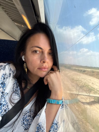 A dark-haired woman with light skin, wearing a white blouse with blue floral patterns and a black crossbody strap, is captured in a selfie taken from inside a train. She has on white wireless earbuds and a gold geometric earring in her left ear. Her right hand, adorned with two gold rings and a turquoise bracelet, rests near her chin. The train's interior is visible in the upper left, with a yellow light panel. Outside the window to the right, a dry, arid landscape of fields and dirt paths is visible, with a bridge crossing over a dry riverbed. The sky is blue with scattered clouds. This scene is taking place in Rosh Haayin, Israel.