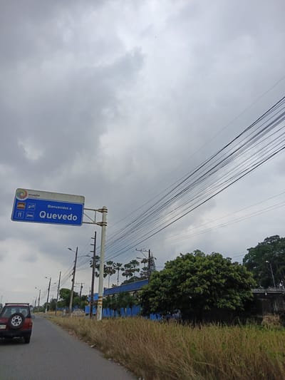 The image captures an overcast day on a road leading into Quevedo, Ecuador, as indicated by a blue and white roadside sign that reads "Bienvenidos a Quevedo". The sign is attached to a metal post and displays the Ecuadorian flag and symbols for restaurants and lodging. Overhead, a network of power lines stretches across the sky. A red SUV is driving on the asphalt road, heading away from the viewer. The roadside is covered with dry, brown grass and vegetation. Further down the road, palm trees and a blue building are visible. Streetlights and utility poles line the road. The sky is filled with grey clouds.