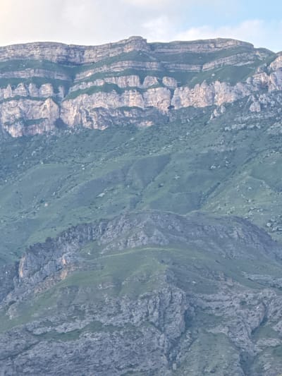 The image displays a panoramic view of mountainous terrain with stratified rock formations and green vegetation. The foreground features rocky slopes interspersed with patches of grass, while the midground shows more rounded, vegetated hillsides. The background is dominated by a large mountain range with distinct horizontal layers of rock, varying in color from light tan to gray, and streaks of green vegetation growing in the crevices. The sky above is a pale blue with some white clouds. The scene appears to be a natural landscape, likely within the region of Khadzhalmakhi, Russia, known for its mountainous geography. There are no people or man-made objects visible in the image, suggesting a remote or natural setting.
