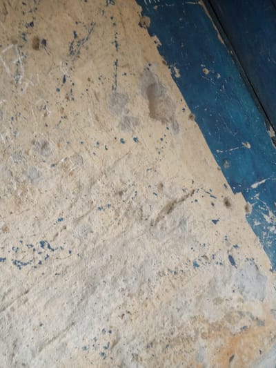 This media file depicts a close-up view of a textured, weathered surface, likely a wall or floor, in Gindin Dutse, Nigeria. The primary subject is the surface itself, which is predominantly a light, sandy beige color, heavily marked with patches of gray and what appears to be old, chipped paint. There are numerous small, dark blue splatters scattered across the surface, suggesting paint or some other liquid stain. Some areas show signs of rougher texture, with raised or chipped sections.
On the right side of the frame, a section of dark, painted wood planks is visible, angled diagonally. This wooden element is painted a deep, vibrant blue, contrasting with the muted tones of the foreground surface. The blue paint also shows signs of wear and tear, with some lighter areas and splatters.
There are no visible people or clear indications of a specific activity or event taking place. The scene appears static and focuses on the materiality and condition of the surfaces. The lighting is somewhat diffused, suggesting it could be daylight, but no distinct shadows or sun position cues are present. The overall impression is one of age and exposure to the elements. The texture and the presence of scattered debris and markings on the surface could imply an outdoor setting or an area subject to wear and tear, such as a public space, a workshop, or a well-used structure. There is no visible text in the image.