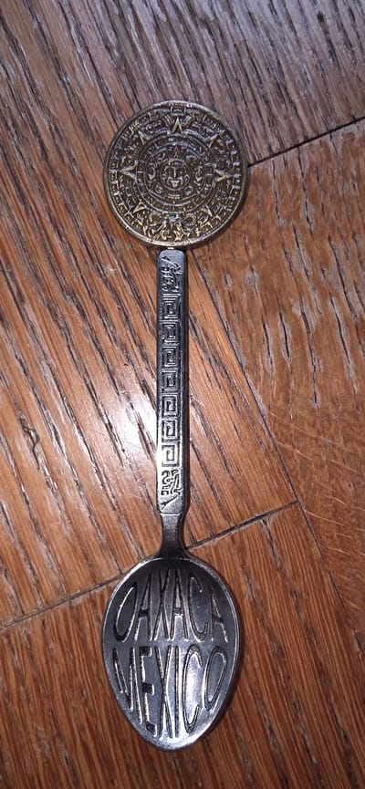 A silver spoon with a decorative coin on the end rests on a wooden surface. The coin is round and features an intricate design, possibly depicting a calendar or sun symbol, with a golden hue. The spoon's handle has a textured pattern of interconnected squares and a small, etched figure near the bowl. The bowl of the spoon is engraved with the words "OAXACA MEXICO" in large, stylized letters. The wooden surface appears to be made of planks with a visible grain and some wear. There are no people or animals present in the image, and the environment appears to be indoors. The image does not provide any information about the city of Marum, Netherlands.