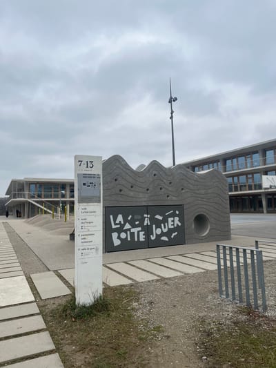 The image captures an outdoor urban area in Meyrin, Switzerland, under an overcast sky. The primary subjects include a distinctive, wave-patterned concrete structure, a tall white directional sign, and modern buildings in the background.

The central concrete structure features textured, undulating surfaces and a prominent dark panel with stylized white text that reads "LA À JOUER BOÎTE," translating to "The Play Box" or "The Toy Box," indicating a space or resource for play. To the left, a tall white sign provides information. Its heading shows "7-13," likely an age range, and "C. Aymonier," possibly a name related to the design or location. Below, a list of numbered facilities is visible: "7 école La Voie Lactée" (school), "9 école des Vergers" (school), "11 parascolaire" (after-school care), "aida," "restaurant," and "13 salle de gym" (gymnasium).

In the background, a multi-story building with numerous windows suggests an educational or public complex. A slender pole equipped with what appears to be a weather station or an anemometer stands against the cloudy sky. The ground is composed of light-colored paving slabs, gravel, and small patches of grass. The scene is static with no people visible, suggesting a quiet moment during the day. The cloudy weather indicates a diffused light, not direct sunlight.