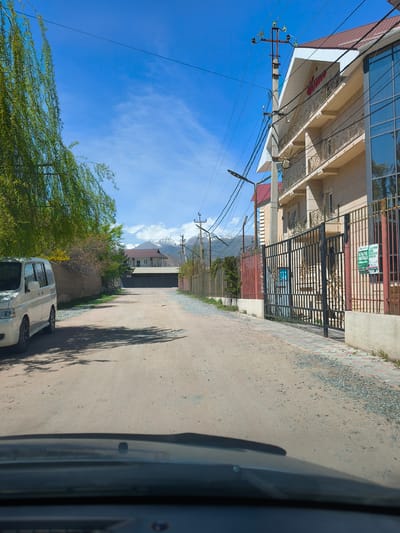 The image depicts a dirt road in the city of Bosteri, Kyrgyzstan, as seen from inside a vehicle. On the left, a light-colored minivan is parked along a wall partially obscured by a willow tree. The road extends into the distance towards a set of dark gates. In the background, mountains with snow-covered peaks are visible. On the right, a two-story building with a dark red sign on the roof, likely a business establishment, is enclosed by a wrought-iron fence. Power lines and utility poles run along the road. The sky is blue with some scattered clouds. The road itself is unpaved and covered with gravel and dust.