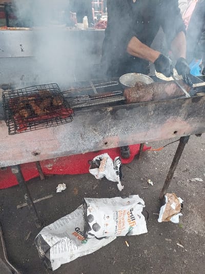 This image depicts an outdoor grilling operation in Eckbolsheim, France. The central focus is a rustic, elongated metal grill with a reddish-brown front panel, elevated on dark metal legs. Heavy smoke emanates from the grill area.
A person, visible from the mid-torso up, stands behind the grill, wearing a dark, long-sleeved garment and black gloves. This individual is actively engaged in cutting or handling a large, elongated piece of dark brown meat directly on the grill surface with a knife or similar tool. An aluminum foil pan is positioned nearby on the grill. To the left, a black metal grate holds several smaller pieces of meat and multiple red sausages. Various metal grilling utensils are also present on the grill surface.
The ground beneath the grill is dark, appearing to be asphalt or dirt. A red mat or carpet is visible directly under the grill. Prominently displayed on the ground is a torn white bag labeled "Grill profi Grillkohle Holzkohle GRILLETTES CHARCOAL GRILL," indicating the fuel source. Several crumpled pieces of paper and other small debris, including cigarette butts, are scattered around the bag and on the ground. The dark-shoed feet of the person grilling are visible near the red mat. The background, obscured by smoke, shows faint outlines of structures or other stalls and indistinct figures.