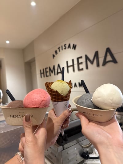 The image shows three individuals holding ice cream treats in front of a wall sign reading "ARTISAN HEMA HEMA ICE CREAM." The ice cream treats are in different flavors: one is a scoop of white ice cream on top of a dark gray scoop in a cardboard container, another is a green scoop on top of a pink scoop in a waffle cone, and the third one is a pink scoop on top of a brown scoop in a cardboard container. Each container has the brand name "HEMA HEMA" printed on it. The individuals are identifiable by their hands and manicured nails. One individual has a bracelet on their wrist. The setting is likely a retail location, an ice cream shop, in George Town, Malaysia.