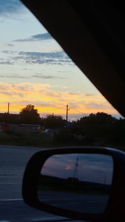 A car's side mirror reflects a dimly lit landscape, possibly in Razeni, Moldova, during twilight. In the reflection, a silhouette of a tower or industrial structure is visible against a darkening sky, with hints of soft pink and orange hues from the setting sun. The foreground of the mirror shows a dark, indistinct horizon line. Outside the mirror, the image captures a sliver of a colorful sunset sky with scattered clouds, transitioning from pale blue to vibrant orange and yellow. Silhouetted trees and a few buildings are visible beneath the sky, and power lines stretch across the scene, supported by utility poles. The overall atmosphere is that of a tranquil evening drive.