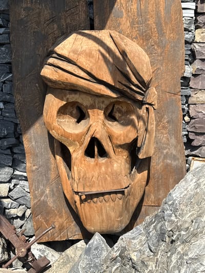 A carved wooden skull with a bandana is mounted on a wooden plank, which is leaning against a stone wall. The skull is carved with eye sockets, a nose cavity, and a grinning mouth with visible teeth. The bandana is depicted as wrapped around the skull's head. The wooden plank shows grain and some rough texture. The stone wall is made of irregularly shaped grey and reddish-brown stones. In the foreground, there are large grey rocks and a rusted metal object resembling an anchor or a propeller with protruding spikes. The overall scene appears to be an outdoor display. This artwork is located at the "Porogi" industrial complex in Satka, Russia, which is known for its outdoor art installations.