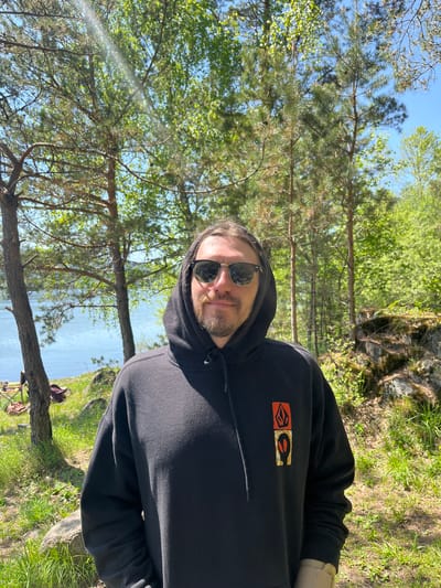 A man wearing a black hoodie with the hood up and sunglasses gazes directly at the camera in a vibrant outdoor setting. He has a beard and his expression is neutral to slightly smiling. On the front of his hoodie, a stylized, multi-colored logo featuring red, orange, and gold shapes is visible. He is positioned near the grassy bank of a calm body of water, likely a lake or wide river, which stretches into the background reflecting the blue sky.
The surroundings are lush with greenery, indicating a natural, forested environment. Tall trees with green foliage and pine needles dominate the background, with a clear blue sky visible through the branches, suggesting bright, sunny weather during the daytime. On the grassy bank in the foreground, what appears to be fishing equipment, including fishing rods on stands and a bag, is set up, implying that fishing is the activity taking place. The scene depicts a peaceful, sunny day in a natural outdoor location, specifically near Sliznevo, Russia.