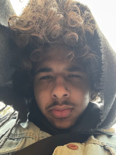 The image depicts a close-up, low-angle selfie of a single male individual, reportedly taken in Meistratzheim, France. The subject, centrally framed, possesses dark, curly brown hair emerging from a dark hooded garment which obscures the upper sides of his head. He has dark eyes, a straight nose, full lips, a light mustache, and minimal chin stubble. He maintains a direct gaze towards the camera. He is wearing a light blue denim jacket, visible across his shoulders and chest, with a copper-toned button on the lower right quadrant of the image. A dark strap, consistent with a vehicle seatbelt, diagonally traverses his upper torso. The background is indistinct and brightly lit, suggesting an outdoor or brightly illuminated indoor environment, with diffuse light illuminating the subject. No specific geographic features of Meistratzheim are discernible within the visible frame.