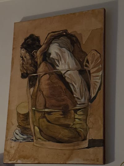 An oil painting portrays a person carrying a bundled load on their back, supported by a woven carrier. The carrier includes a large, fan-like leaf atop the load. Predominantly brown and white hues define the subject, with visible brushstrokes suggesting a rough texture. The lower portion features a support structure, possibly a resting platform, adding depth to the scene. A signature is discreetly placed in the upper right corner.