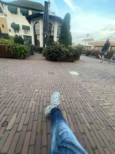 The image depicts an outdoor scene in Oud-Beijerland, Netherlands. The ground is paved with red bricks, and a person wearing jeans and grey Balenciaga sneakers is visible in the lower right of the frame, appearing to be seated. Adjacent to the brick pathway, there are large planters containing greenery, including shrubs and small trees. In the background, buildings with white facades and green awnings can be seen. Further into the background, other people are walking along a paved area with outdoor seating. The sky is partially visible and appears overcast.