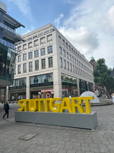A low-angle, full outdoor shot shows a large, bright yellow "STUTTGART" sign in the foreground, resting on a gray rectangular base. The sign is positioned on a paved pedestrian area, which appears to be a city square. Behind the sign, a modern building with a beige facade and numerous windows dominates the mid-ground. Below the windows on the ground floor, large storefronts are visible, including "RESERVED" and "POP UP OUTLET" signs. Several bicycles are parked on the left side of the image. Further in the background, a fountain with water spraying upwards is partially visible. To the right of the fountain, trees and a building with a green dome can be seen. The sky above is partly cloudy. A few people are visible in the scene, including a person walking away from the camera on the left, and others strolling in the background near the fountain and shops. The overall impression is that of a lively urban environment.
