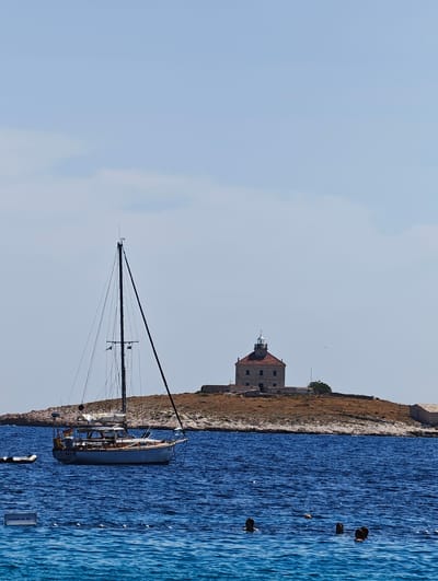 A sailboat is anchored in the blue waters near a small island with a lighthouse and some stone buildings. The lighthouse is made of stone with a red roof and a lantern room. Several people are swimming in the water in the foreground. The sky is a clear blue with a few wispy clouds. The island appears to be rocky and covered in dry grass. A small inflatable dinghy is tied to the stern of the sailboat. A signpost is visible in the water with the text "visithvar.hr".