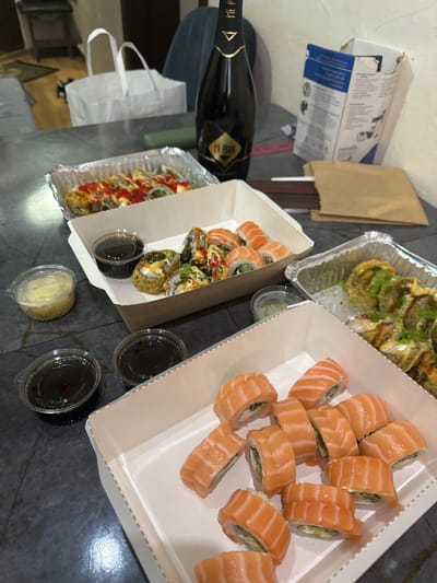 The image depicts a selection of sushi takeout containers on a dark gray surface. Three containers are visible: one rectangular container filled with salmon sushi, another with a mix of salmon sushi and fried sushi rolls, and a third containing what appears to be fully fried sushi rolls with various toppings. Condiments, including soy sauce and ginger, are also visible in small plastic containers. A dark-colored bottle, possibly wine or sparkling cider, lies near the sushi containers, along with a discarded cardboard box and a brown paper bag. The environment appears to be indoors, likely a dining table or countertop. There is no indication of the specific location in the city of Sîngera, Moldova, and no people are visible in the image. The focus is on the food arrangement and accompanying items.