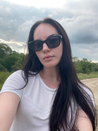 The image depicts a selfie-style portrait of a woman, taken outdoors in the city of Zlokuchene, Bulgaria. The subject, positioned centrally, has long, dark brown hair cascading over her shoulders and chest. She wears dark, rectangular-framed sunglasses with reflective lenses, obscuring her eyes, and a white, short-sleeved t-shirt with subtle shimmering details. Her lips are naturally colored and slightly parted.
The background features an overcast sky filled with grey and white clouds, indicating diffused lighting. Below the horizon line, dense green foliage of trees and shrubs is visible, forming a vegetated landscape. The immediate ground behind the woman is an unpaved dirt path with sparse green vegetation. The overall setting is a natural, outdoor environment under an overcast sky.