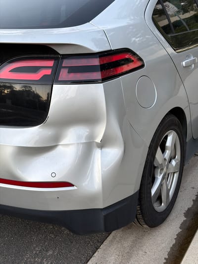 A silver Chevrolet Volt is parked on a street in Aliso Viejo, United States of America. The rear passenger-side quarter panel of the vehicle has been significantly damaged with a large dent. The dent spans from the taillight downwards towards the bumper. The car is a dark silver color with black trim around the windows and a black rear valence. The rear driver-side taillight is visible and appears undamaged. A portion of the rear passenger-side wheel, a silver alloy rim with five spokes, is also visible. The car is parked on asphalt pavement, with a concrete curb to the right. The sky, reflected in the car's body, indicates it is daytime.