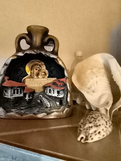 The image depicts a close-up shot of several decorative objects placed on a shelf. In the foreground, to the left, is a ceramic vessel shaped like a hollowed-out urn with two loop handles at the top. The opening of the urn reveals a miniature diorama featuring traditional houses with red roofs and arched windows, a stone bridge, and an icon of the Virgin Mary holding the infant Jesus. The icon is placed on what appears to be pages from a book. To the right of the urn, there are two seashells, one large and spiraled, and a smaller, speckled shell in front. Behind the shells, a blurred object, possibly a plastic bottle, is visible. The background is a plain, textured wall. The lighting is soft, casting gentle shadows. There are no people visible in the image, and no specific location in the city of Žiaukos, Lithuania, is indicated.