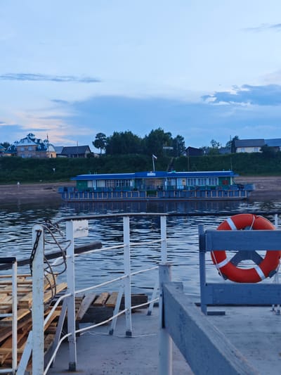 This media file depicts a serene river scene at dusk in Vorogovo, Russia. The foreground is dominated by a ferry or barge, with a weathered deck, metal railings painted light blue, and a prominent orange life preserver with white stripes attached to a support structure. Piles of wooden pallets are stacked on the left side of the deck, suggesting cargo or supplies.
In the middle ground, a long, narrow boat with a green hull and a blue roof is docked or moored near the shore. It appears to be a passenger vessel or a ferry, with numerous windows running along its length. The water is calm, reflecting the soft, diffused light of the approaching evening.
The background features a verdant riverbank lined with trees. Beyond the trees, several houses and buildings are visible, with some displaying pitched roofs. One building in the distance appears to be a large, light-colored structure with dark blue accents. A flag flies from a pole near one of the buildings. A sign with Cyrillic text, "ВОРОГОВО", is visible on the bank, confirming the location.
The sky is a soft blue with wispy clouds, indicating a tranquil evening sky after sunset. The overall mood is peaceful and quiet, evoking a sense of calm and the end of a day. The time of day is clearly dusk, with the low light casting a gentle glow over the scene. The weather appears to be clear and calm.