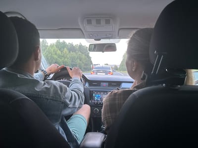 Inside a car on a road in Verkhnechusovskie Gorodki, Russia, a person drives with their hands on the steering wheel, wearing a denim jacket and shorts. To the right, another person sits in the passenger seat, facing forward and wearing a plaid shirt. The car's dashboard displays a media screen. Through the windshield, trees line the road, and another car is visible ahead.