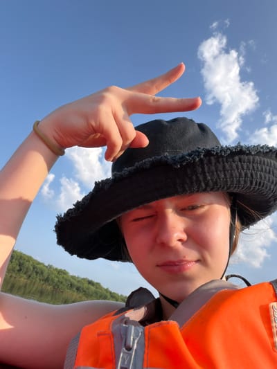 A young person wearing a black bucket hat and an orange life vest is taking a selfie. They are holding up their hand in a peace sign gesture. The background shows a blue sky with white clouds and a green, tree-lined body of water, suggesting an outdoor activity likely on a lake or river in Kazaevo, Russia. The sun is casting a warm glow on the person's face.