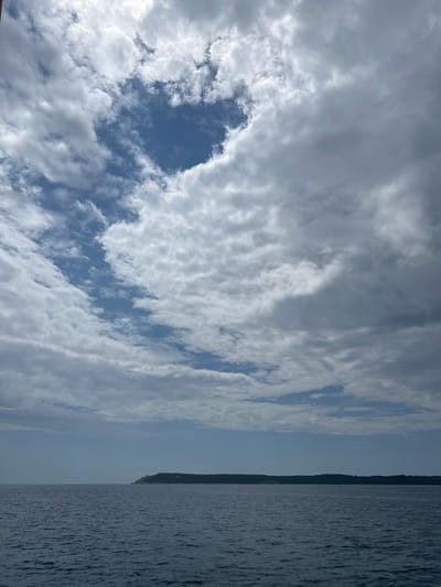 The image shows a body of water adjacent to a line of land. The water surface is textured with small ripples, reflecting light. The land appears to be covered with trees. The sky above is filled with clouds of varying density. Blue sky is visible in patches between the clouds. The image has been rotated 90 degrees counter-clockwise. The location is assumed to be in or near Luštica, Montenegro, but there are no features in the image which confirms this, and there are no people, objects, or actions visible in the frame.