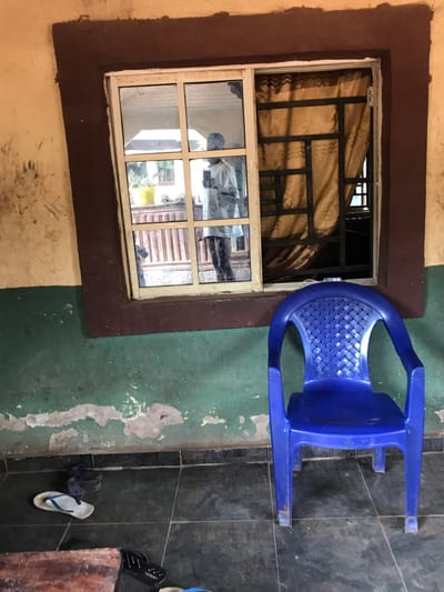 A window with brown trim has metal bars and a tan curtain partially obscuring the view. The window reflects objects from the outside. Next to the window sits a blue plastic chair. The wall is painted two colors: green at the bottom with peeling paint, and yellow at the top. Two pairs of sandals and a pair of flip-flops are on the tiled floor.