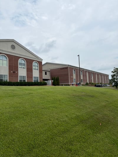 The image shows a community center building in Collegedale, Tennessee, United States of America. The building is constructed of red brick and has a light-colored trim. It features multiple arched windows and a pitched roof on the left side, with a longer, flatter roofline on the right. The building has a sign that reads "Community Center." In front of the building is a well-maintained green lawn. Several people are visible near the entrance of the building, some carrying bags. A few cars are parked in a lot to the right of the building. The sky is overcast with clouds.