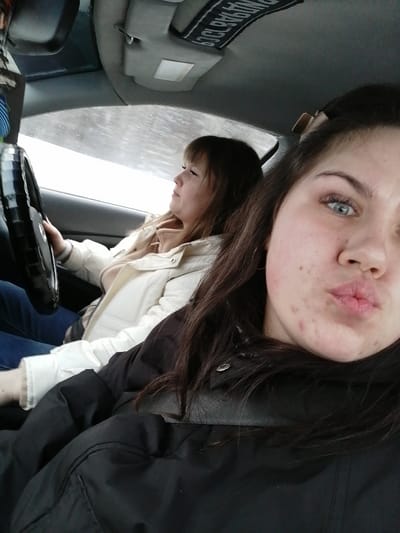 The image shows two young women inside a car, with one in the passenger seat taking a selfie.
In the immediate foreground, a young woman with long, dark hair and light blue eyes is looking directly at the camera. Her fair skin shows some visible acne or blemishes on her cheeks and chin. She has a playful expression, with her lips pursed in a slight pout. She is wearing a black puffer jacket, and her dark hair frames her face and falls over her shoulders.
Behind her, in the driver's seat, is another young woman. She has straight, light brown hair with bangs, and she is looking forward, presumably at the road ahead. She is dressed in a light-colored, possibly cream or off-white, quilted or puffer jacket over a lighter top, and dark blue jeans are visible on her lap. Her hands are on the steering wheel, which is covered with a black, textured wrap.
The interior of the car is visible, including parts of the dashboard and the ceiling. A sun visor is flipped up on the passenger side, and another is visible above the driver's head. On the driver's sun visor, there is a black fabric strap or tag with white text. Although appearing upside down and slightly distorted, the word "DURABILITY" is clearly discernible, and below it, a four-digit number that resembles "1989" or "1969" can be faintly made out. The view through the windshield is bright but hazy or diffused, suggesting either an overcast day, rain, or snow, indicating it is daytime. The women's jackets suggest cool or cold weather. The overall scene captures a casual moment during what appears to be a car ride.