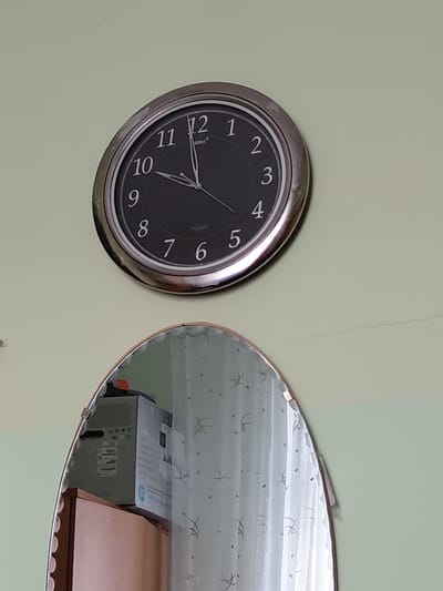 The image presents an indoor scene featuring a wall-mounted analog clock and an adjacent, irregularly shaped mirror. The wall is painted a light, muted green or pale yellow hue, with a subtle vertical crack visible above and to the right of the clock.
The round clock has a silver-colored metallic frame, showing some signs of wear. Its black face features clear white Arabic numerals from 1 to 12. The time displayed by the white hour and minute hands is approximately 9:45, with the hour hand positioned between 9 and 10, and the minute hand directly on the 9. A slender white second hand is also visible. Small white text, likely a brand name "RIFAT", is printed above the 12 numeral.
To the right of the clock, the mirror, with a thin metallic trim, reflects a portion of the room. The reflection clearly shows white or light-colored curtains adorned with a repeating pattern of abstract shapes or possibly stars. Below the curtains, a grey cardboard box, possibly packaging for an electronic device, is visible. It features barcodes and some unreadable text, with a partial "HP" logo discernible on its side. A reddish-brown wooden surface, likely a table or shelf, is partially seen behind and next to the box.
No people are present, and no specific activity is taking place. The scene is static, depicting an ordinary interior setting. No explicit visual cues within the image indicate the provided location context of Nadym, Russia, nor is any specific time of day or weather discernible from the indoor environment.