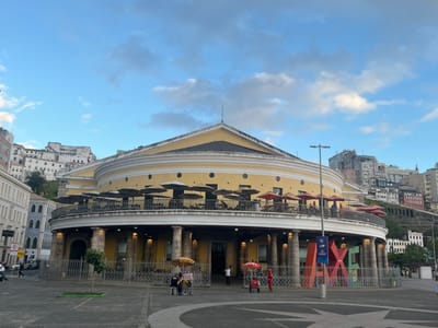 This image displays the Mercado Modelo in Salvador, Bahia, Brazil, a large, two-story, circular building with a distinctive yellow facade. The ground level features an open arcade with columns, enclosed by a dark metal fence. The upper level presents an expansive outdoor terrace, equipped with numerous tables and chairs, largely shaded by an array of black, red, and yellow parasols, indicating an active dining or seating area. A yellow conical roof tops the structure.
In the foreground, a paved plaza exhibits a decorative star-shaped pattern. A prominent, colorful sculptural sign spelling "AXÉ" (with red, blue, and green letters) stands before the building. Two street vendors operate pushcarts with umbrellas on the plaza near the building's entrance; one cart is yellow, the other red, with patrons seated nearby. Several individuals are visible on the plaza and the street to the left.
The background to the left reveals older, multi-story buildings, primarily white and blue, on an incline. To the right, a dense concentration of modern and older multi-story buildings, varying in color and style, ascends a steep hillside. The sky is partially cloudy blue, indicating daylight.