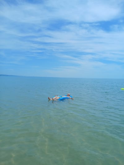 A person lies on a bright blue inflatable raft in the calm, shallow sea off the coast of Kranevo, Bulgaria. The water is clear and translucent, revealing the sandy seabed below. The sky is a brilliant blue, adorned with wispy white clouds. In the distance, a faint, hazy shoreline can be seen. The overall scene conveys a sense of peaceful relaxation on a sunny day.