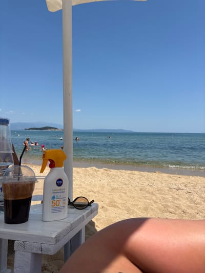 In Ολυμπιάδα, Greece, on a sunny day, a white pole stands on a sandy beach. In the foreground, a white wooden table holds a plastic cup of iced coffee, a bottle of NIVEA SUN sunscreen, and a pair of sunglasses. The ocean is visible in the background with several people swimming. The sky is clear and blue.