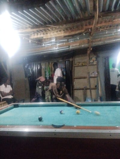 A dimly lit indoor scene features a man playing pool on a green table. He leans over, cue in hand, focusing on the balls. A black ball sits near the left edge of the table. Behind him, onlookers gather, including adults and children. The room's ceiling has a corrugated metal roof. Shelves are visible along the back wall, and a person stands in a doorway in the background.