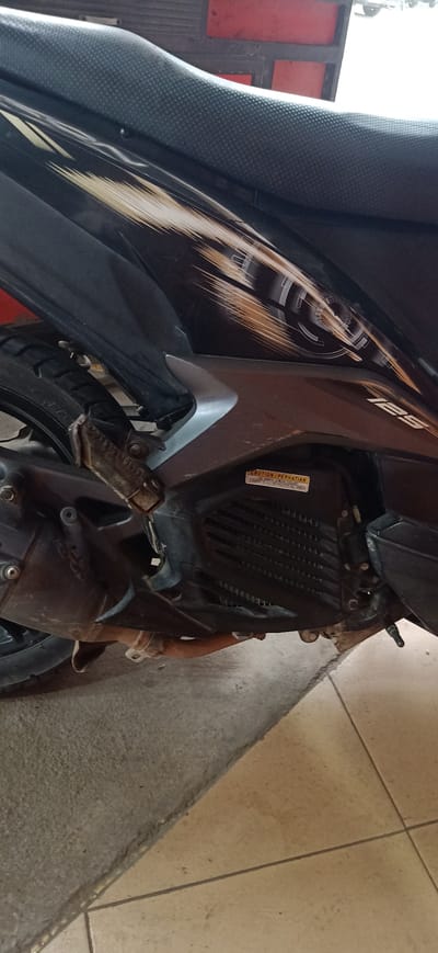 The image shows a close-up view of a motorcycle's side, focusing on its engine and part of the seat. The motorcycle is black with a brown and gold striped decal running along the side panel. The seat is textured black. Below the seat, the engine casing is visible, with a ribbed cover and a warning sticker. The sticker contains text in both English and what appears to be Indonesian. The English text reads "CAUTION | PERHATIAN," and below that, "IMPORTANT: FAILURE TO FOLLOW INSTRUCTIONS CAN RESULT IN SERIOUS INJURY OR DEATH." The Indonesian text appears to be a translation of the English warning.
Visible mechanical parts include the exhaust pipe, which is a rusty brown color, and part of the rear tire. The motorcycle is parked on a surface that transitions from rough concrete to beige tiles. The lighting suggests it is daytime, possibly indoors or in a shaded outdoor area, as there are no strong shadows. The scene appears to be a casual setting, perhaps a garage or a parking area in Gampongteungoh, Indonesia, given the location context. There are no people visible in the image, and the focus is solely on the motorcycle. The overall impression is that of a functional, used vehicle.