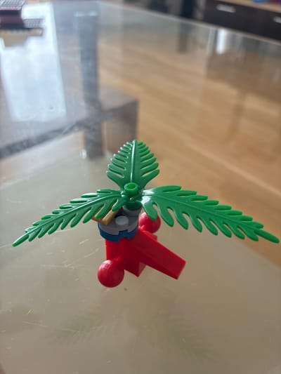 The image shows a close-up, overhead view of a small, red and blue toy airplane with green palm fronds attached to its wings. The toy is placed on a reflective surface, likely a table, which shows a wooden grain pattern in the background. The lighting suggests it could be daytime. The toy appears to be a single object, possibly part of a larger play set, and there are no people visible. The scene is simple and focused entirely on the toy. There is no visible text.