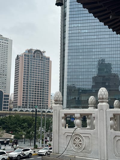 This is a vertical, full shot of a street scene in Jing'an District, China, likely during the day under overcast skies. The foreground features a white stone balustrade with decorative circular motifs and pineapple-like finials. Beyond the balustrade, a modern glass-fronted skyscraper dominates the background, reflecting the sky and surrounding buildings. To the right, several white cars, possibly police vehicles due to their markings, are parked along the street. People are visible near the cars and on the sidewalk, suggesting pedestrian activity. Behind the vehicles, a multi-story building with a grid-like facade is visible, and further back, another building is under construction, with its upper floors still exposed. The scene captures a typical urban environment with a blend of contemporary architecture and signs of everyday life. Some text is visible on the white cars, but it is too small and pixelated to be legible. A street sign is also visible on a pole, but its text is not discernible.