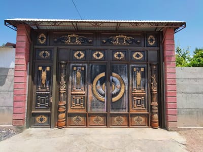 The image shows a large, ornate metal gate in Sovet, Kyrgyzstan. The gate is dark brown with intricate gold detailing and appears to be made of wrought iron. It is divided into multiple panels, with decorative elements such as floral motifs and geometric patterns. The center panels feature a circular design with a concentric ring pattern. The gate is flanked by two brick pillars, one of which is visible on the left side of the image. The gate is partially covered by a metal roof supported by a metal frame. The ground in front of the gate is concrete. In the background, there is a concrete wall and some greenery. The sky is clear and blue. The image is taken in daylight.