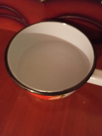 An overhead, close-up, slightly blurry shot shows the interior of a white enamel mug filled with a pale liquid, possibly water, or a milky beverage. The mug has a dark rim, and the exterior is partially visible, with a red surface showing some floral detailing. The mug rests on a red surface, possibly a table or countertop. The context of Nayman, Kyrgyzstan, is not discernible from the image itself.